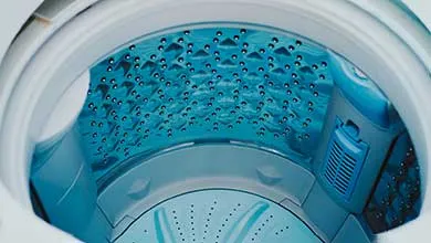 Close-up of an empty, top-loading washing machine drum with blue-tinted, dimpled stainless steel walls and a light blue agitator at the bottom.