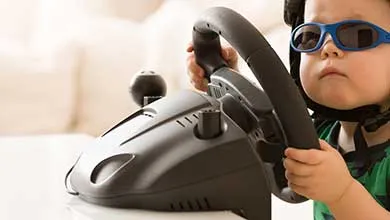 Child in helmet and blue sunglasses holds a video game steering wheel.