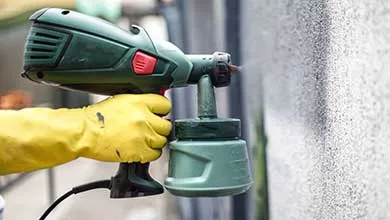 A person wearing a yellow glove uses a green electric paint sprayer to apply paint to a textured gray wall.