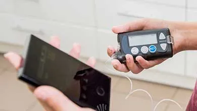 A person holds a black insulin pump with a clear tube in one hand and a smartphone in the other.