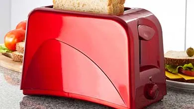A shiny red two-slice toaster on a dark countertop with one slice of bread in a slot.