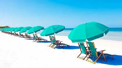 A row of turquoise beach chairs and umbrellas on white sand faces a calm ocean under a clear blue sky.