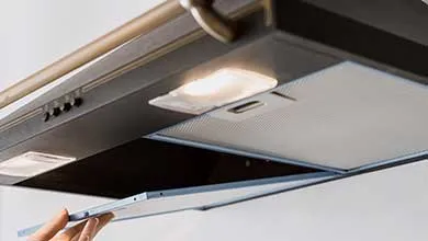 A hand opens a rectangular metal mesh filter on the lit underside of a kitchen range hood.