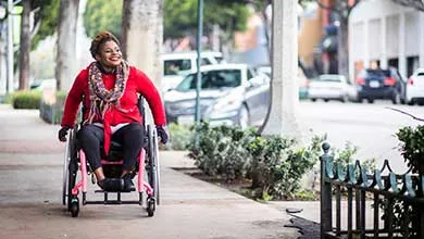 Smiling Black woman in a red sweater and scarf using a pink wheelchair on a city sidewalk.
