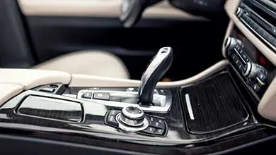 Close-up of a luxury car’s center console with an upright automatic gear shift, glossy black trim, metallic buttons, and beige leather upholstery. Part of the dashboard climate controls is visible.